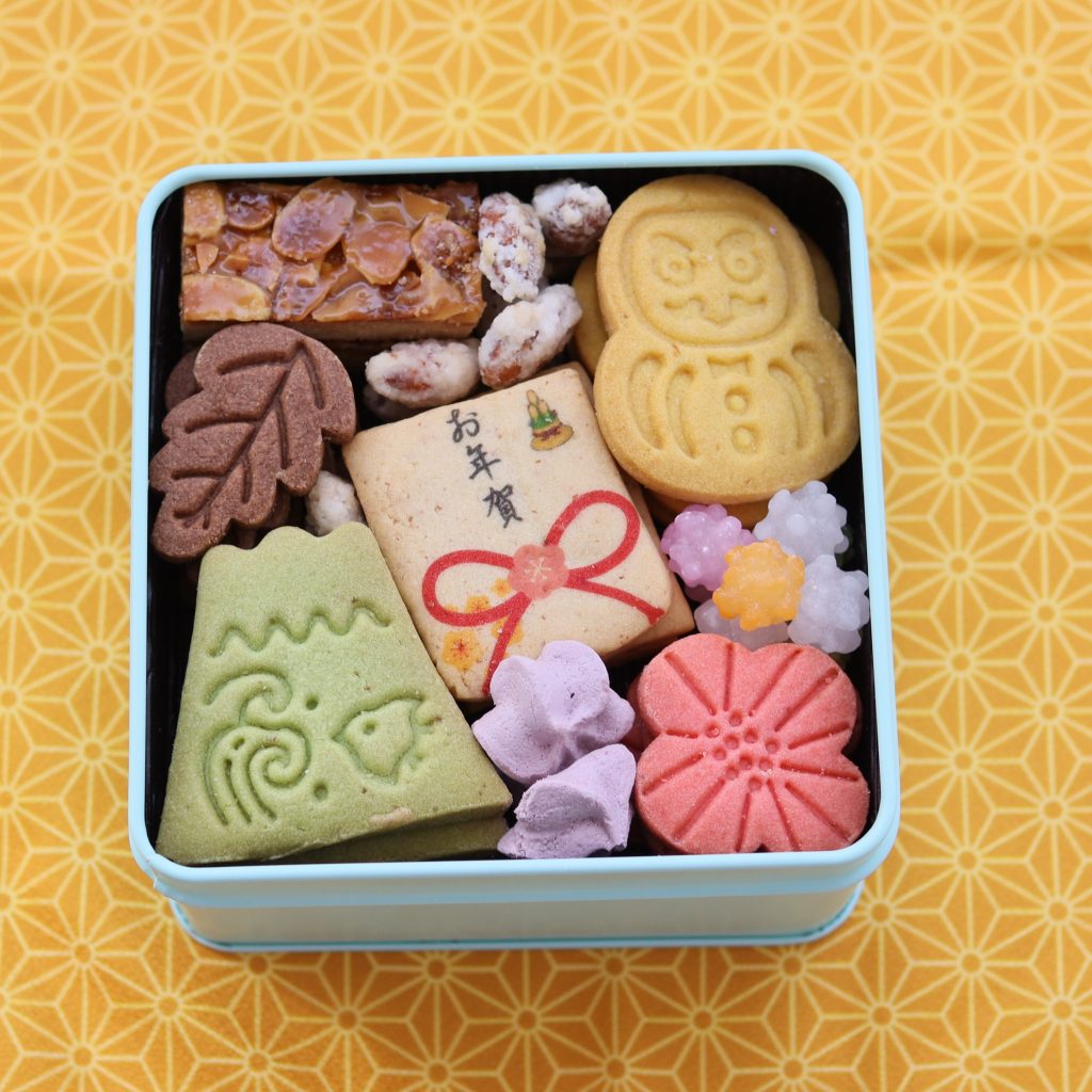 お正月モチーフのクッキーが詰まったカラフルな菓子缶。干支、富士山、花などの形が並ぶ。