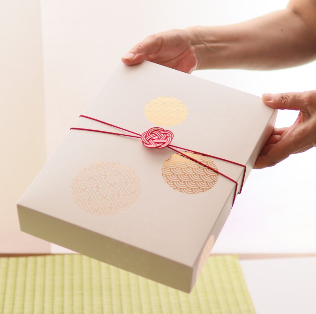金色の和柄模様があしらわれた白い化粧箱を手渡ししている様子。贈り物のイメージ。